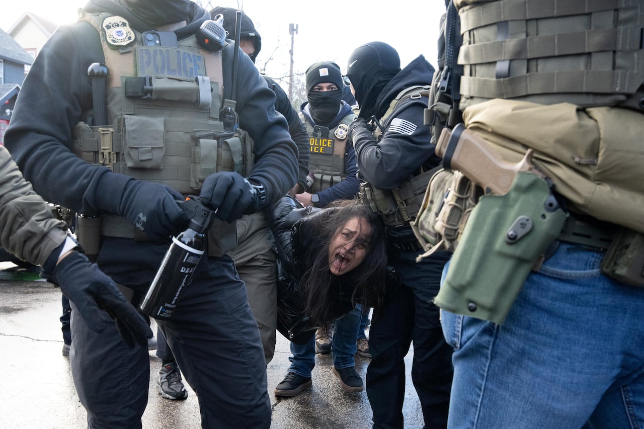 Several masked people wearing police vests hold a woman in a parka, her long, dark hair partially covering her face as she yells.