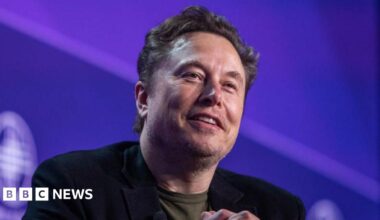 Elon Musk stares towards the camera with a smile while sitting against a purple background on a stage