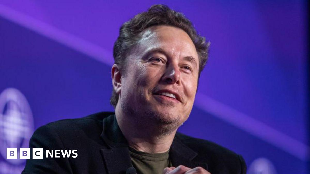 Elon Musk stares towards the camera with a smile while sitting against a purple background on a stage