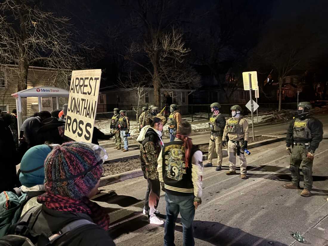Demonstrators stand facing front federal immigration agents who are masked and wearing helmets. One demonstrator holds a large sign that reads, "Arrest Jonathan Ross."