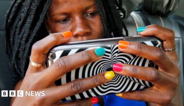 A woman with braids and multi-coloured nail varnish hold ups a mobile phone encased in a zebra design cover