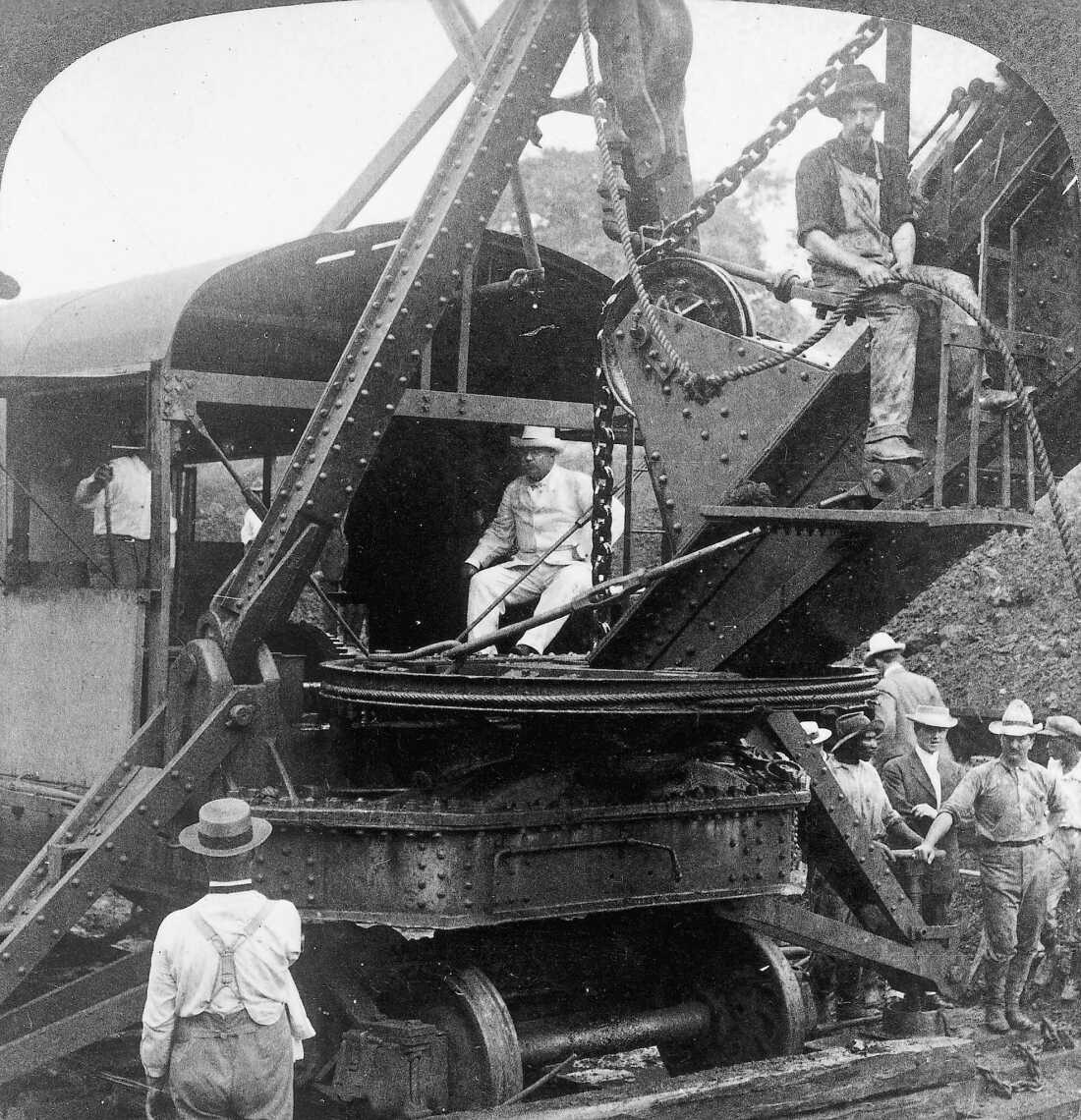 In this black-and-white photo, President Theodore Roosevelt sits on a steam shovel at the Culebra Cut of the Panama Canal in November 1906. He's wearing a light-colored suit and hat. Multiple workers stand around the large excavating machine.