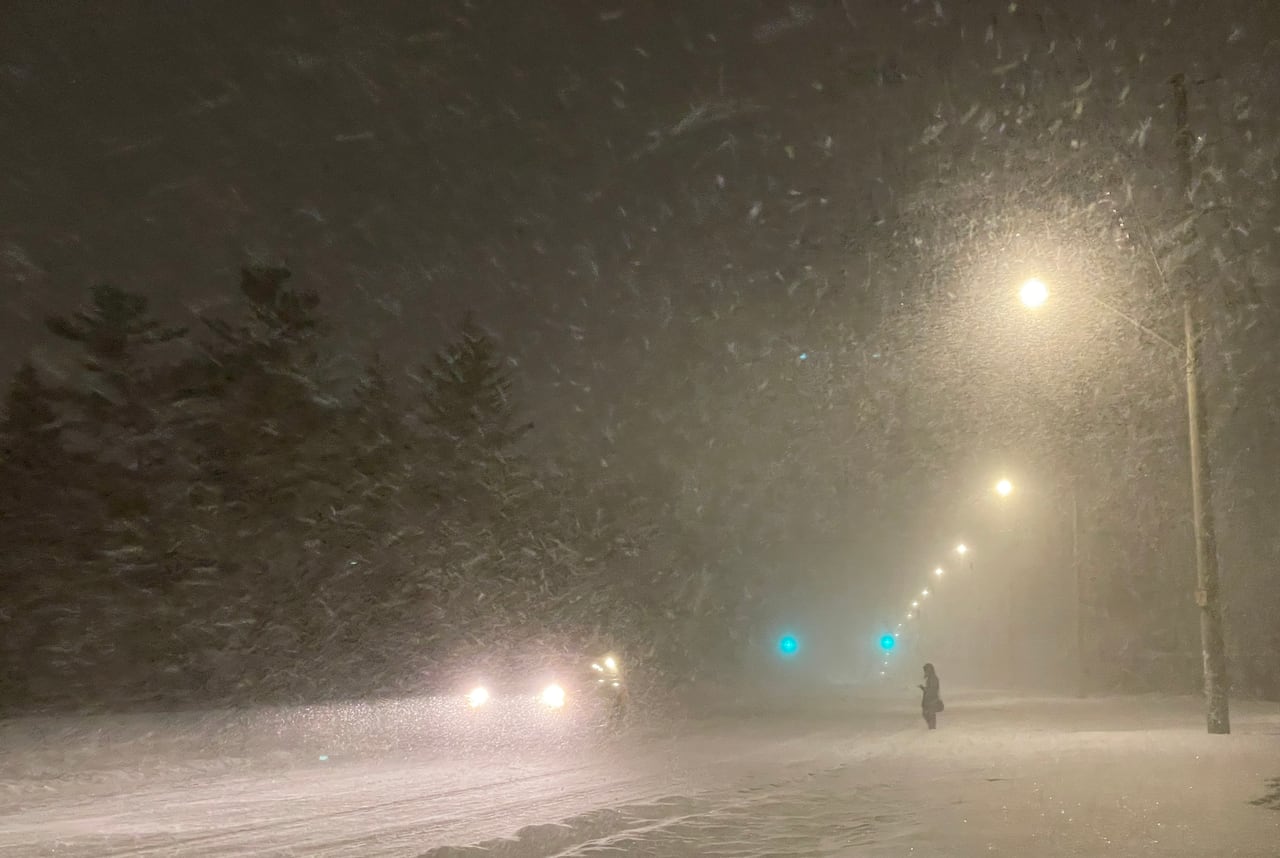 A person stands on a snow-covered street as a car drives by. It's dark out and the snow is still falling