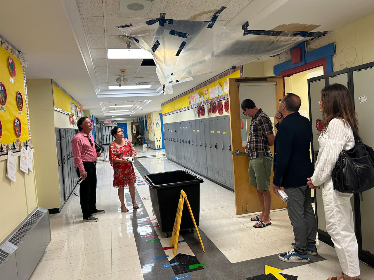 Three men and two women stand in a school hallway, looking up at plastic sheeting taped to the ceiling above a large black bin.