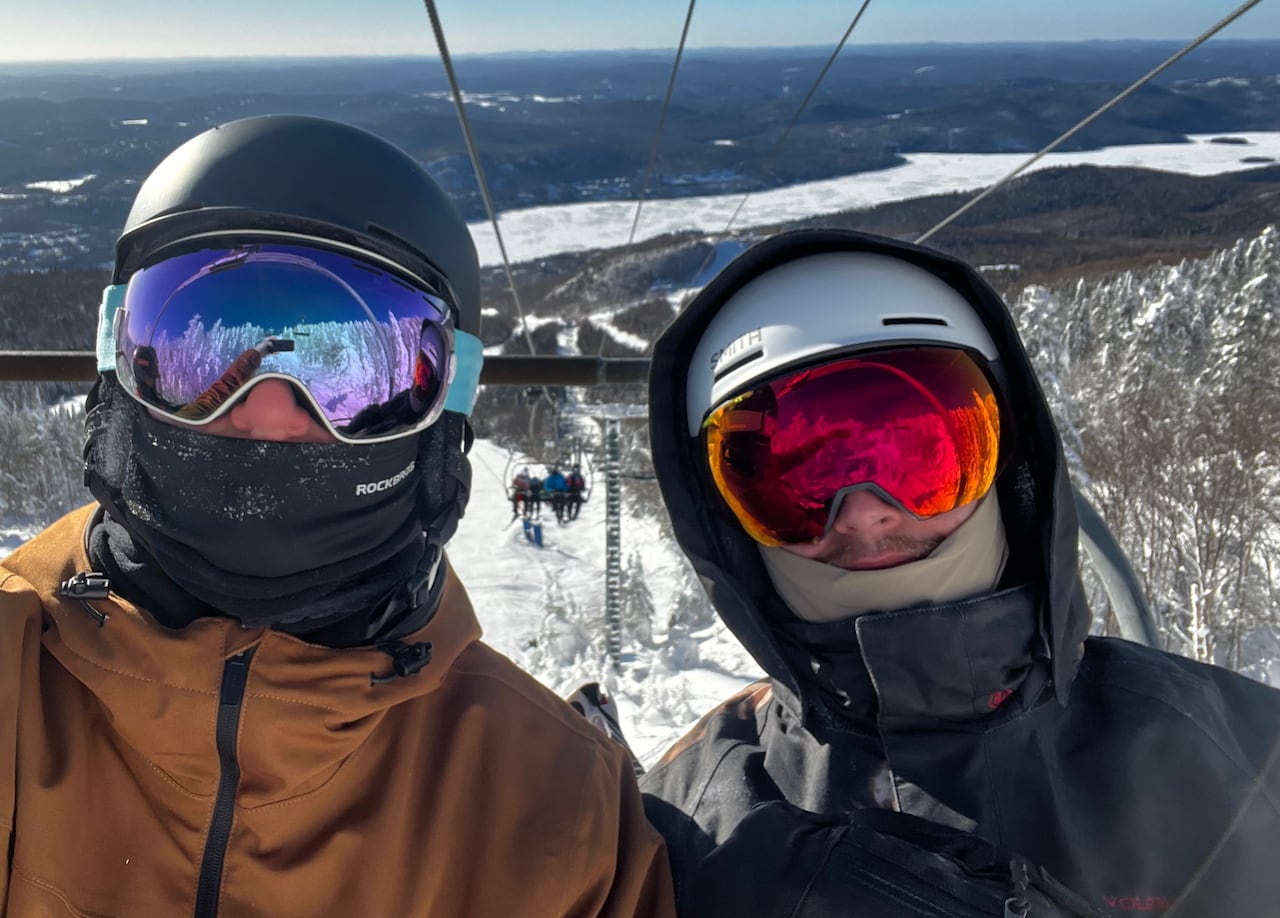 Two people in ski helmets and goggles taking a selfie while riding a chair lift