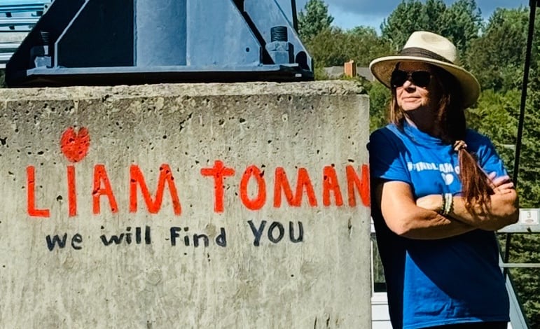 A woman leans against a concrete pillar with the words "Liam Toman we will find you" painted on it.