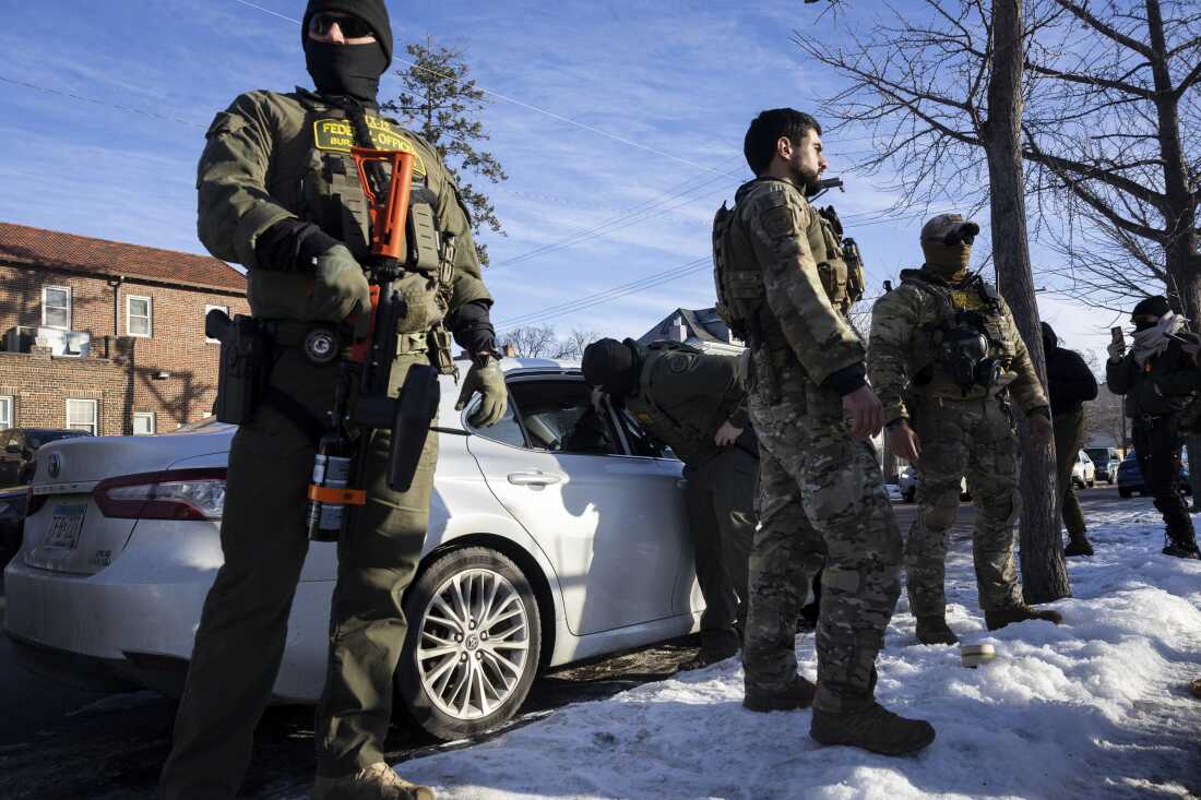 Customs and Border Protection officers stand near a car parked at the curb. 