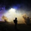 Clouds and bursts of tear gas fill a nighttime scene with two law enforcement officers standing in silhouette.