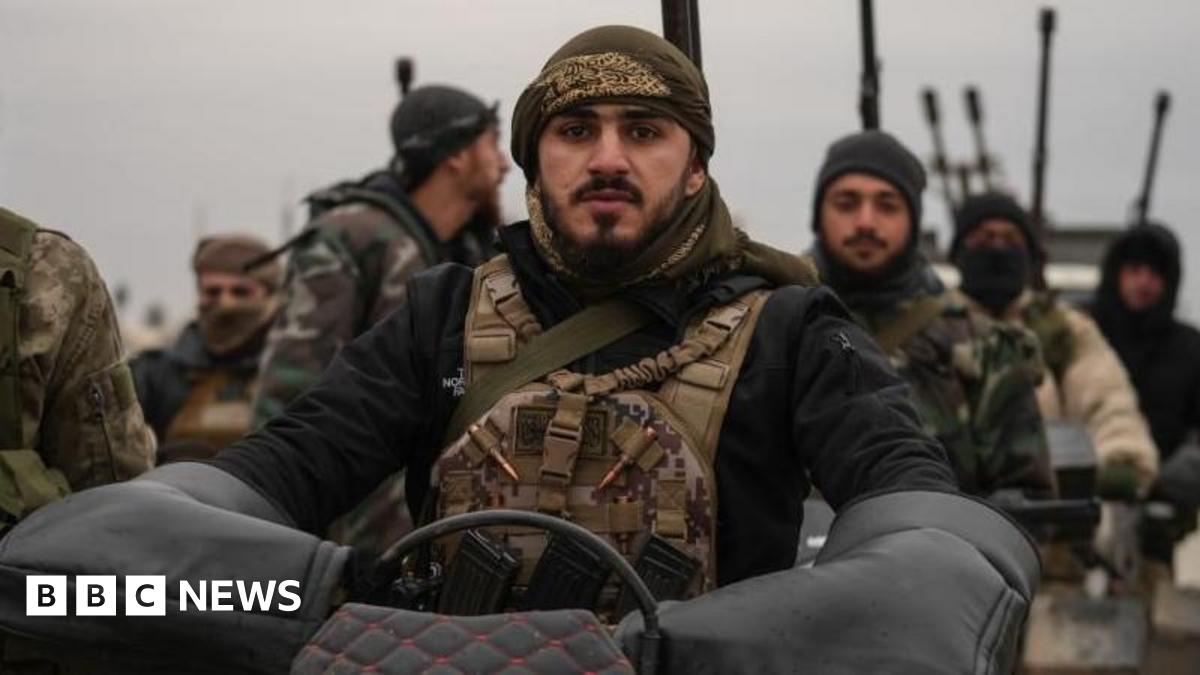 Syrian troops stand together as one soldier sits on a motorcycle, wearing tactical gear and surrounded by others carrying weapons.