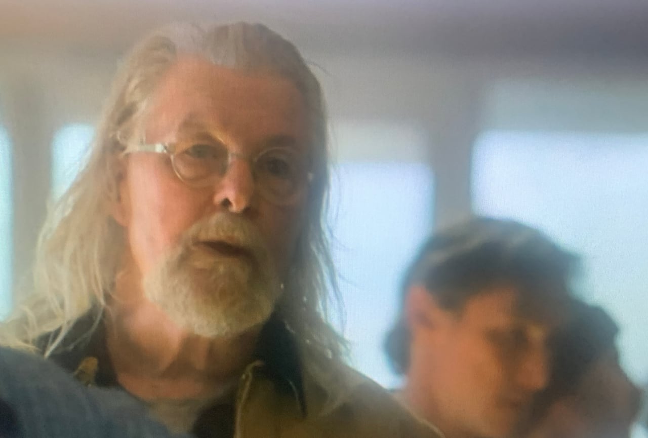 An older man with glasses, white beard and tan jacket stares forward