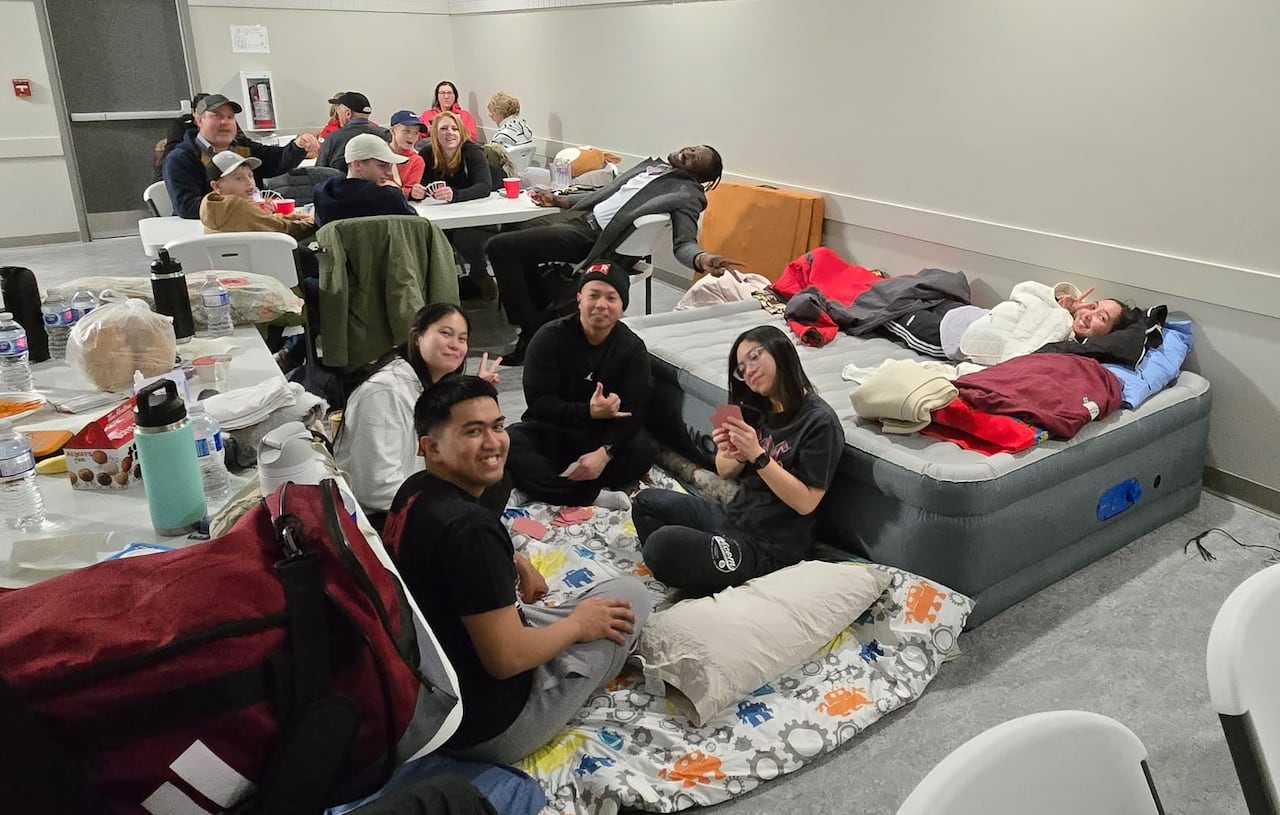 A group of people are in a room, some sitting on the floor playing cards, others on a table and a person is laying on a mattress.
