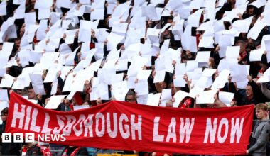 People stand in front of the Hillsborough Memorial outside Anfield Stadium in Liverpool. There are flowers and heart balloons in front of a plaque with the names of the 96 victims of the disaster.