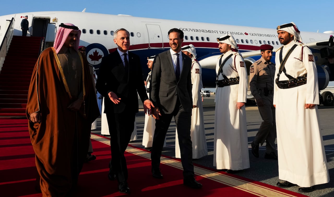 Three men walks on a red carpet on a tarmac with a plane behind them.