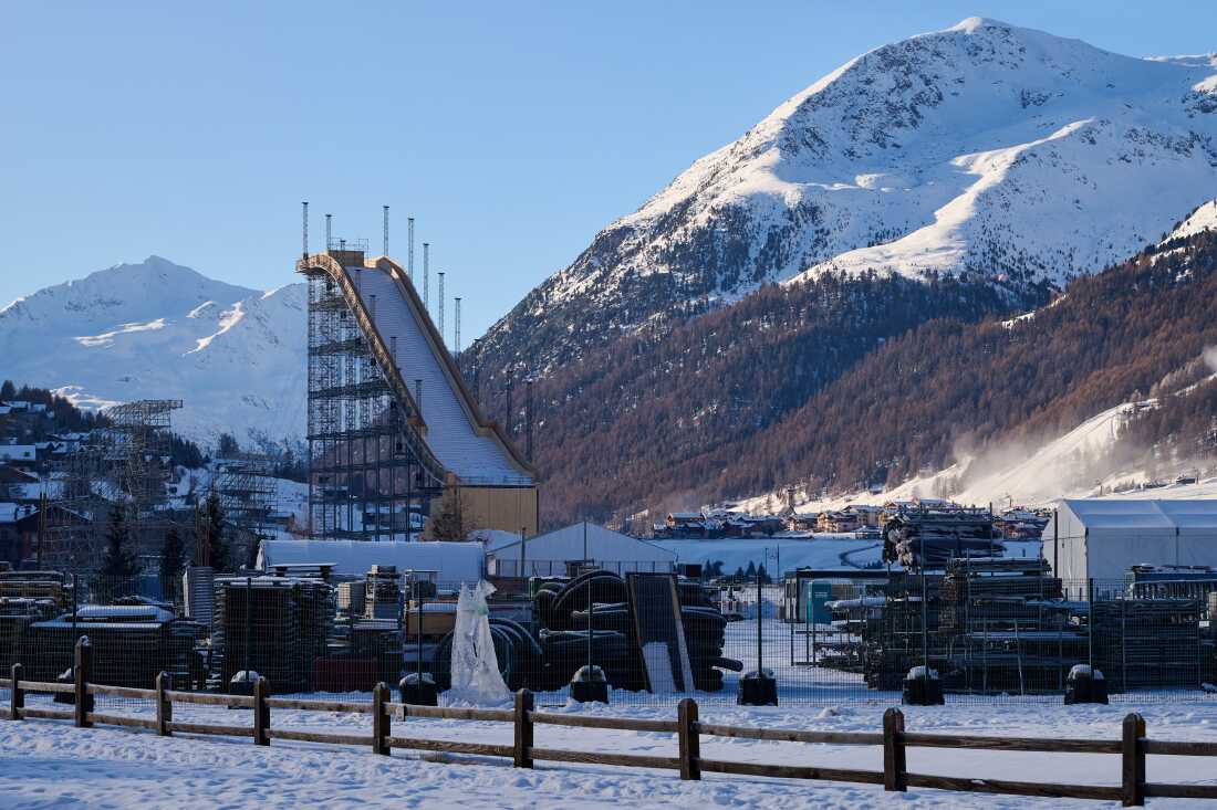 A steep, long slope is shown mid-construction in Livigno Snow Park in December. The bottom part of the slope has yet to be constructed. Peaks covered by snow and Alpine trees are in the background.