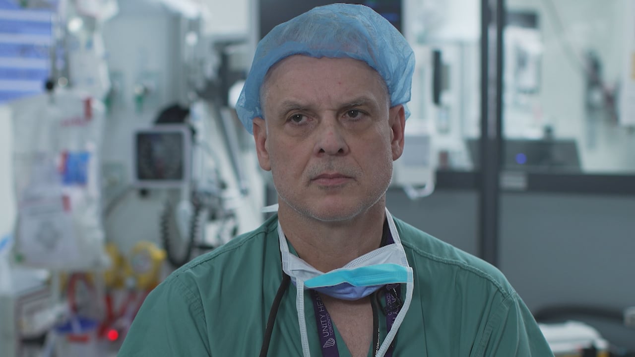A man wearing hospital scrubs, a blue hair cap and a mask hanging around his neck looks off-camera. 