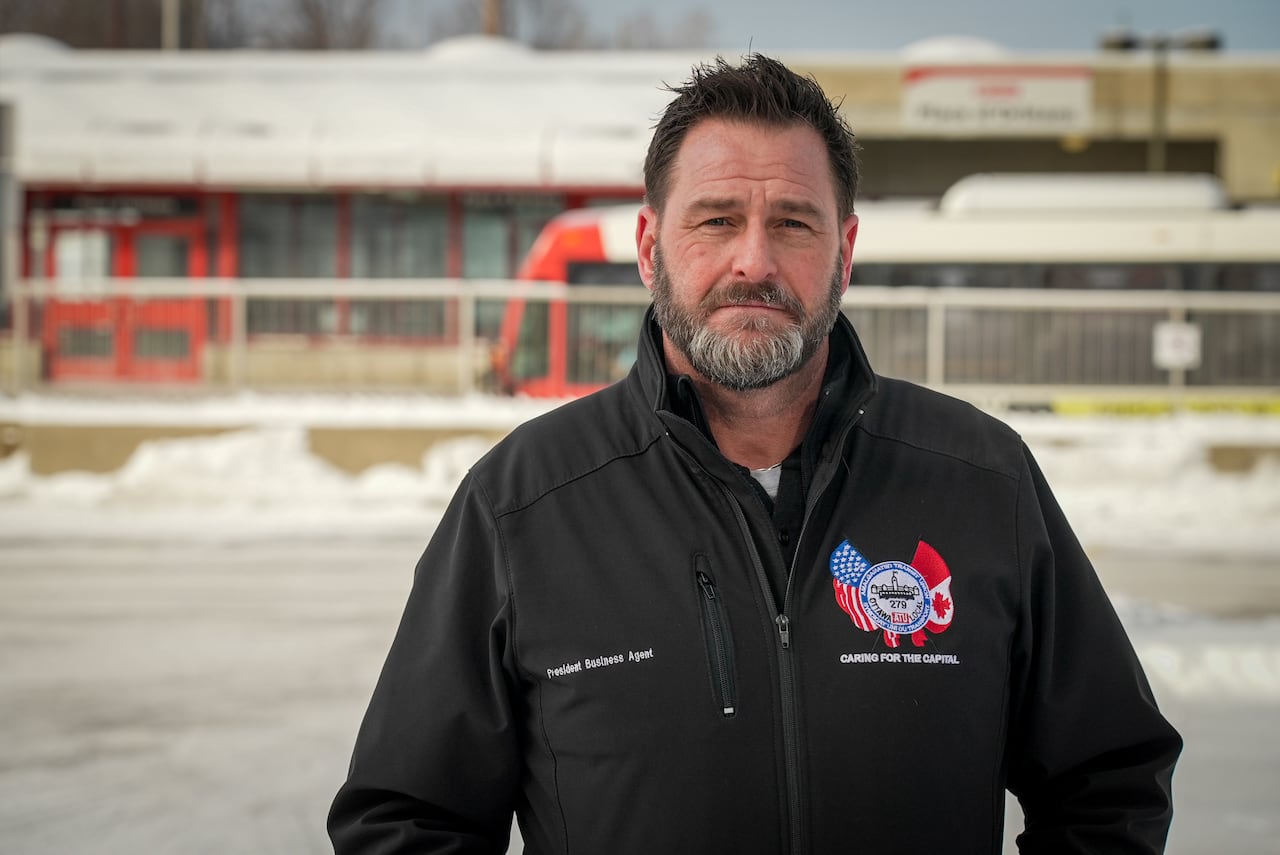 A bearded man with an embroidered union jacket in front of a bus stop.