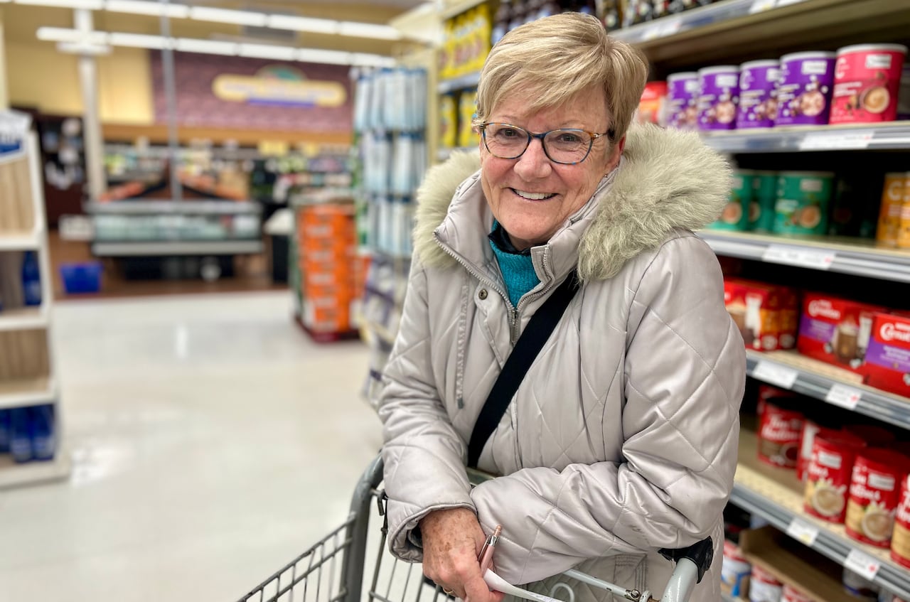 A woman with short, blond hair and a grey winter coat smiles at the camera while leaning on her shopping cart in a grocery store. 
