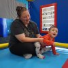 A woman sits on a padded floor mat while playing with her toddler. Her hair is pulled back and her sunglasses are pushed back on her head.