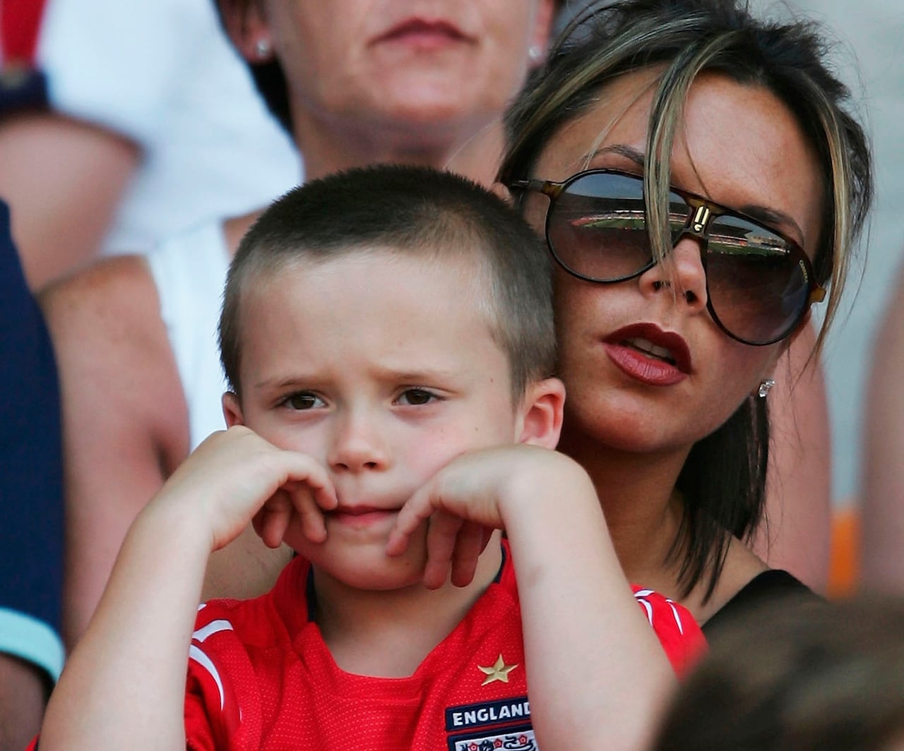 A woman with short brown hair, with blonde tips, and wearing sunglasses, holds a young boy, whose head is shaved and wearing a red soccer jersey with a gold star and and England badge on it.