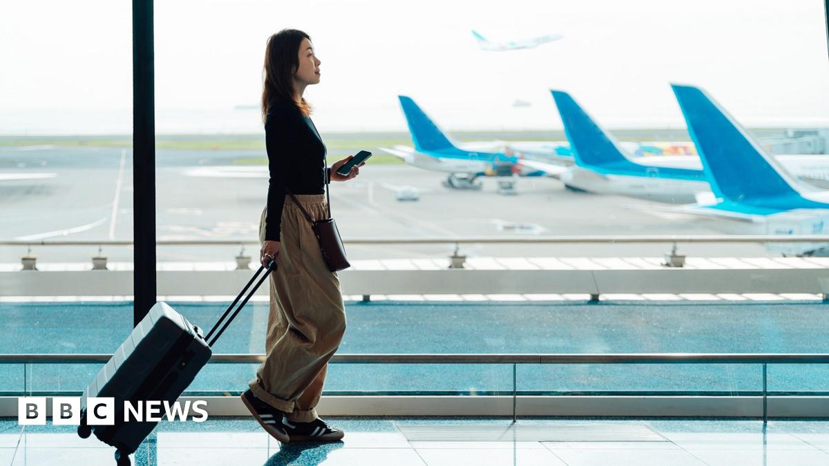 A young woman walks in an airport with her carry-on luggage. She is carrying her phone and wearing baggy beige trousers and a black top