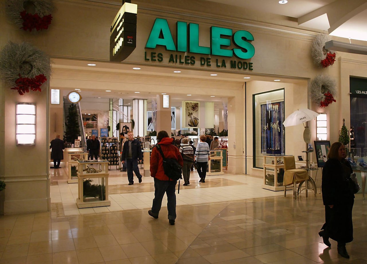 People walking by a Les Ailes de la Mode location inside a shopping mall. The shop has a bright green sign 