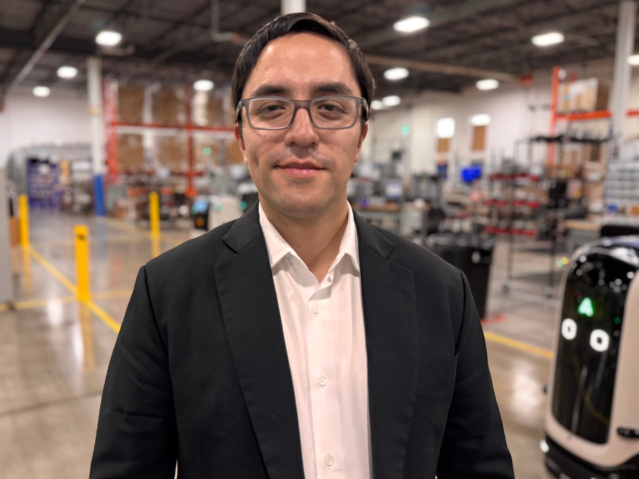 A man with glasses and brown hair stands in a factory and smiles at the camera