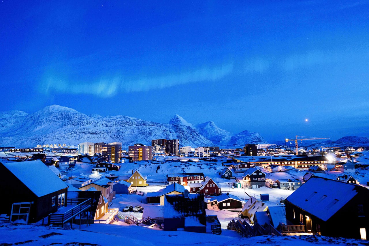 A small city is seen in front of snowy mountains and below a dark blue sky with a white aurora borealis.