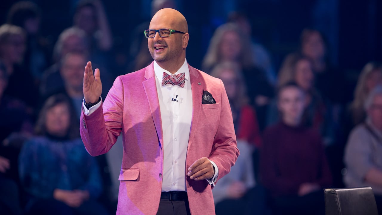 A bald man in a pink suit jacket gestures towards a studio audience.