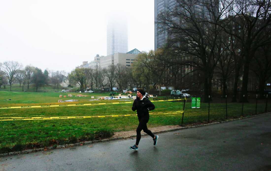 A jogger is seen in warm winter running clothes on a paved path in front of a wide open, green lawn. Buildings are in the background.