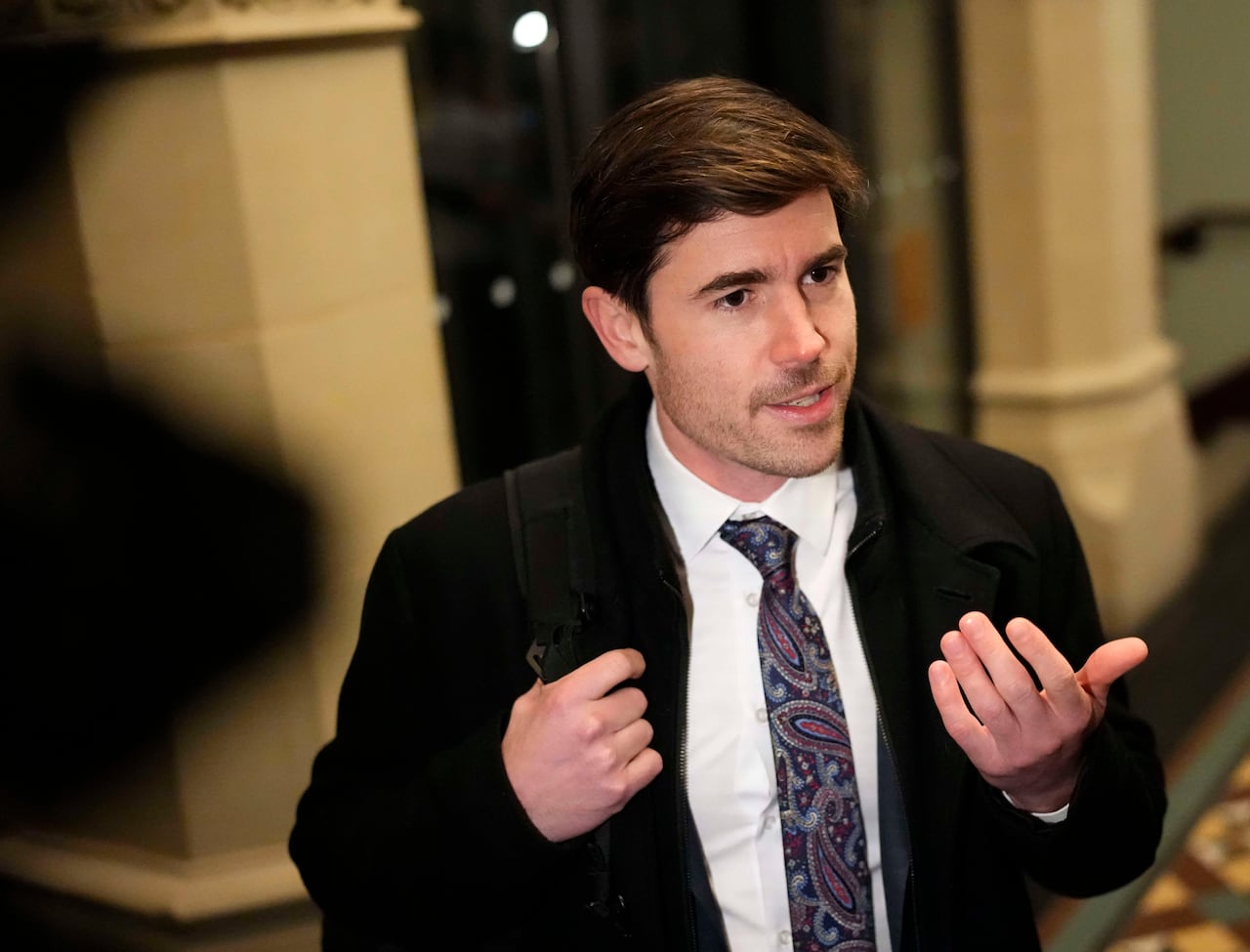 A white man wearing a patterned tie and suit speaks in a corridor.