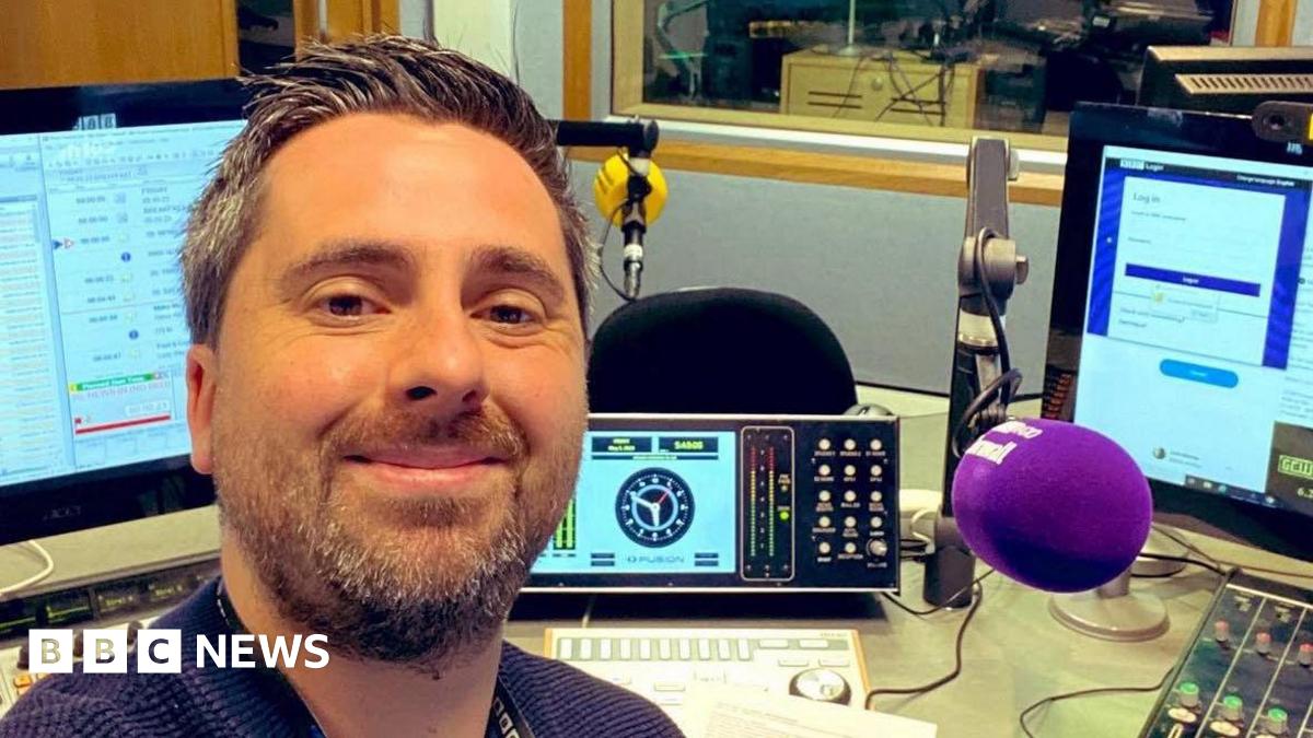 Jack Murley pictured taking a selfie in a Radio studio. He is smiling at the camera. He is sitting at a desk and in the background there are various recording deviscs, controls , a microphone and computer screens.