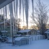 Icicles hang down from the eaves of the roof in the foreground of the photo, looking out onto a snowy neighborhood landscape, including a backyard and surrounding houses, during a winter storm. 