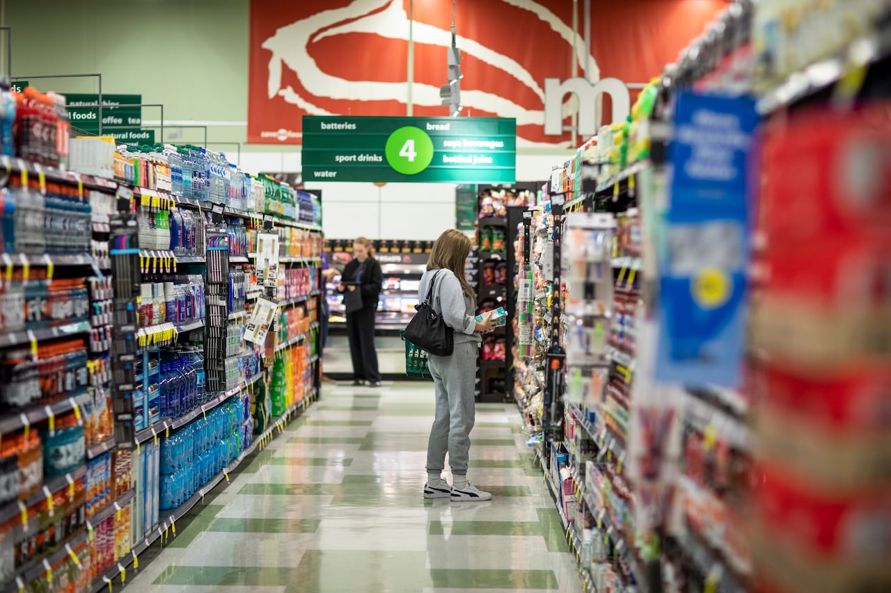 A woman in grey track pants looks at an item in a grocery store aisle.