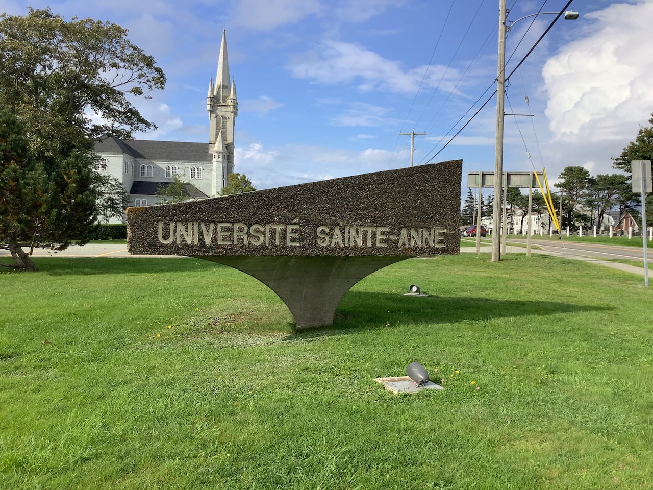 A sign situated on grass says Université Sainte-Anne with a church and a streetscape in the background.