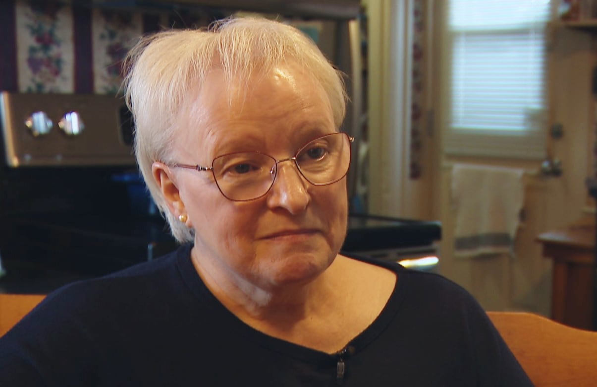 A woman with short white hair and glasses sitting in a kitchen chair, with her hands held together