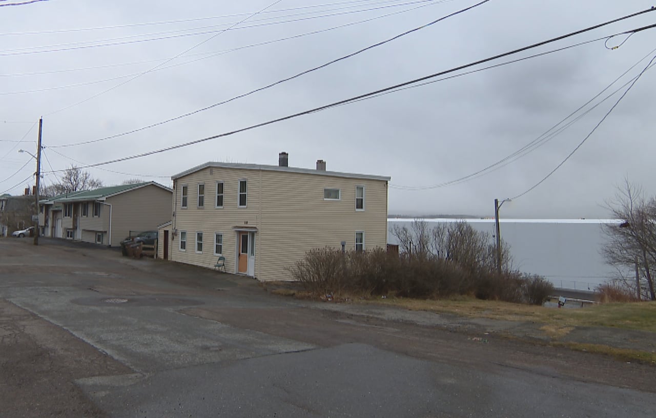 A view of a street with homes along the left side. Behind the homes is a long, white building.