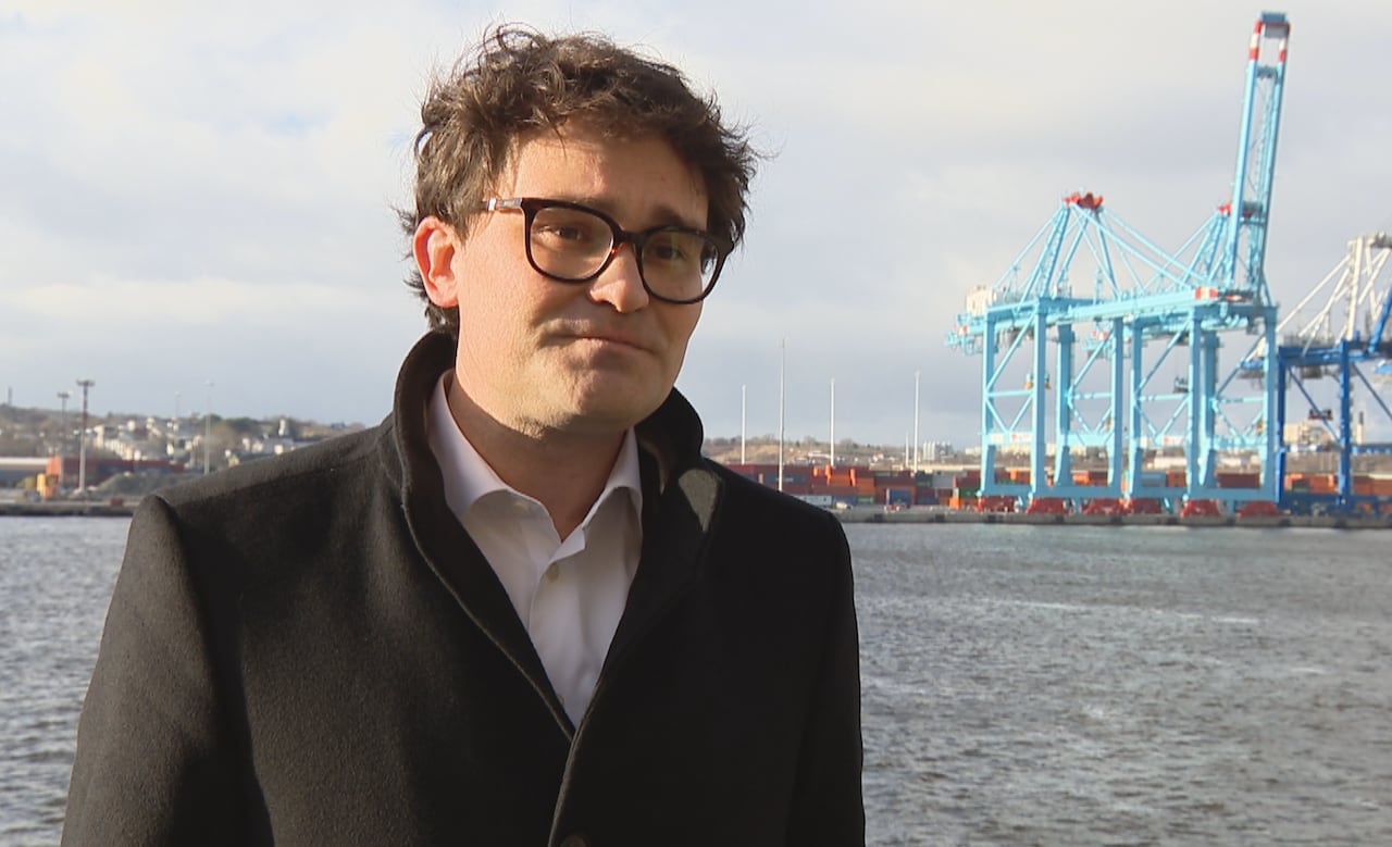 A man with black hair and glasses, wearing a black coat, standing in front of a harbour with cranes and shipping containers in the background.