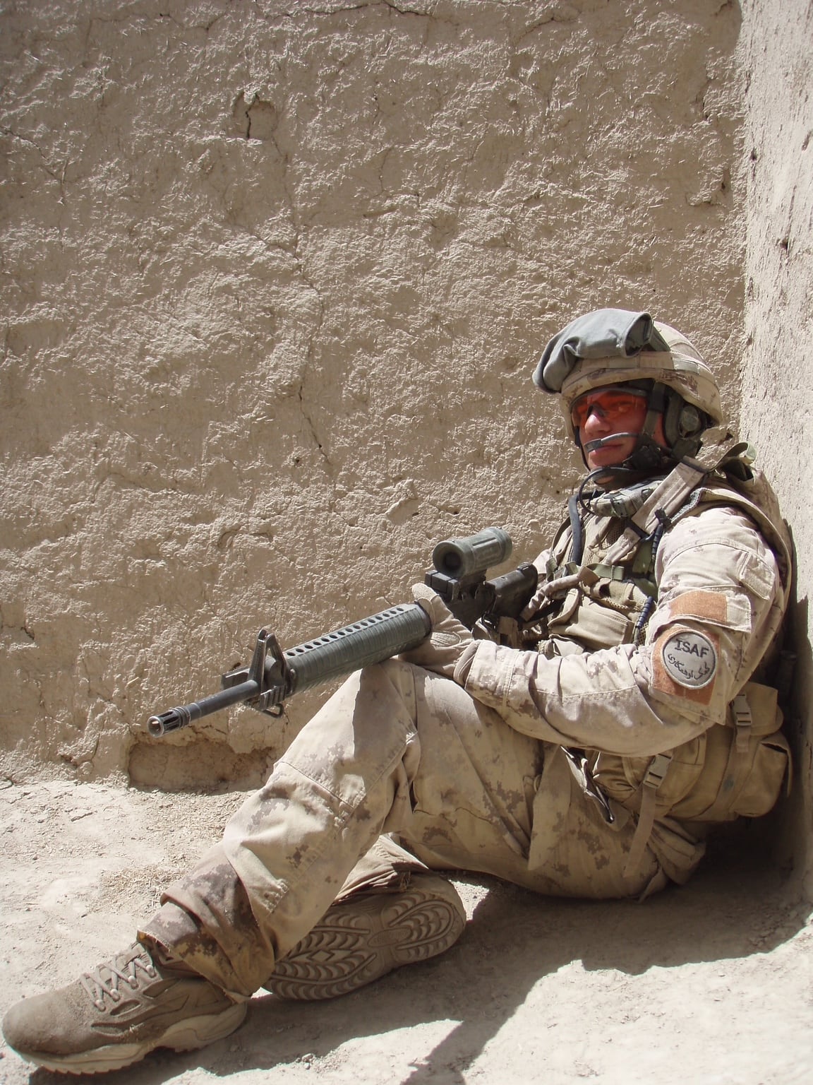 A soldier, holding an assault rifle and wearing desert camoflage, crouches against a wall