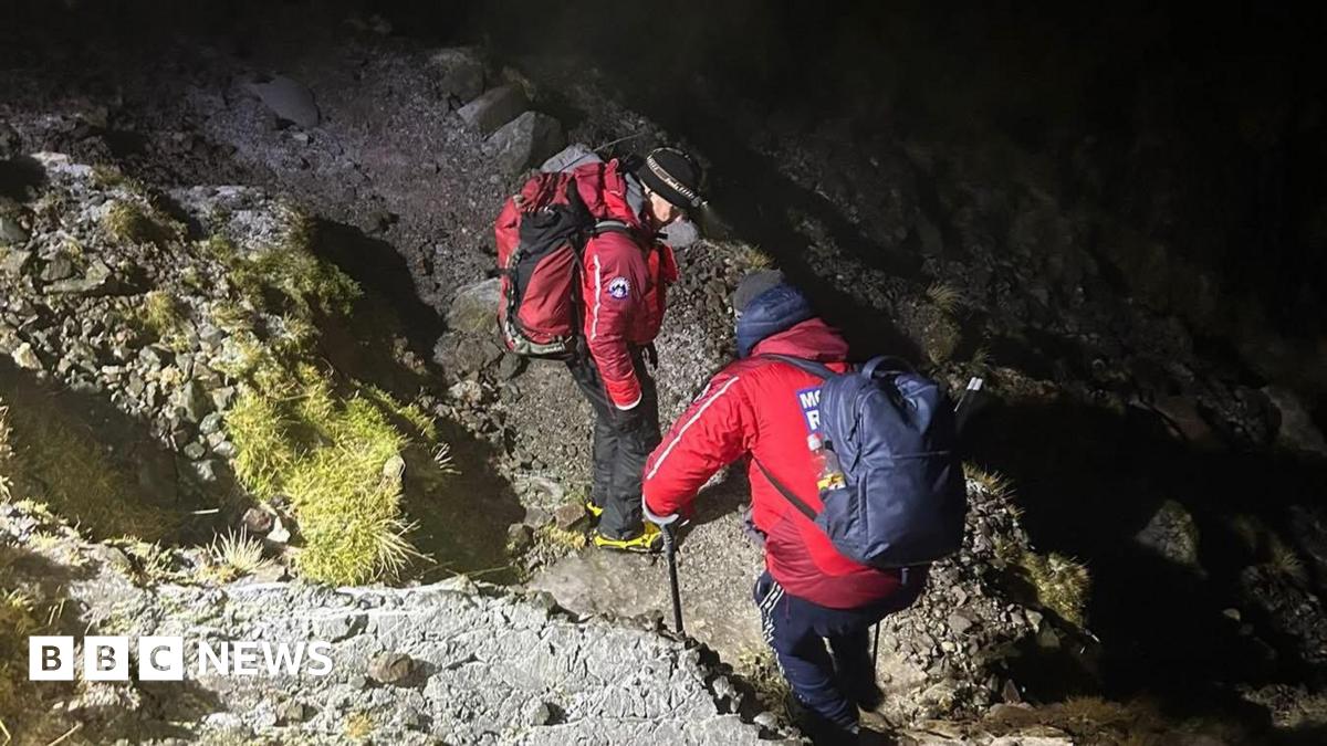 Two mountain rescue volunteers walk on the side of a craggy mountain in the dark, wearing headtorches. They are wearing red mountain rescue branded jackets and carrying rucksacks.