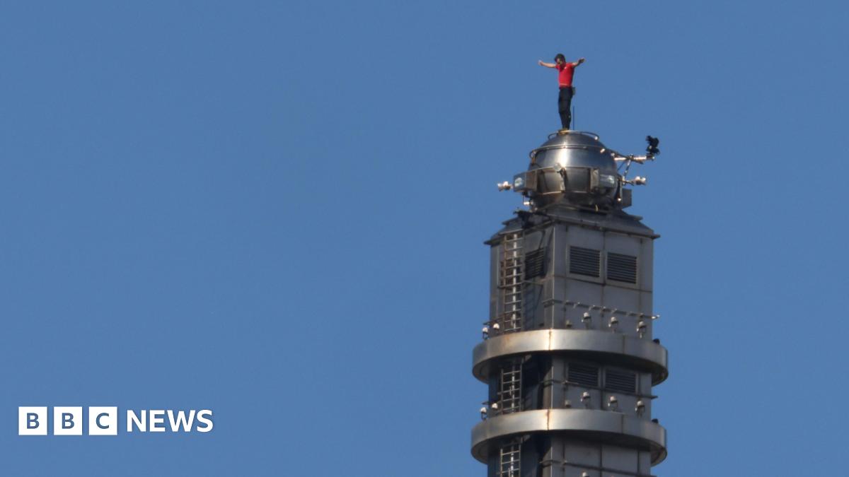 Alex Honnold stands on top of the Taipei 101 skyscraper building in a red t-shirt and dark trousers. There is only blue sky behind him.