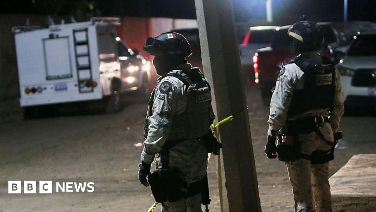 Members of the National Guard stand at the crime scene where at least 11 people were killed and 12 more wounded during an armed attack at a football field in Salamanca, Guanajuato state, Mexico, on January 25, 2026. The two men are wearing helmets and are in uniform. Cars and vans can be seen in the background as well as yellow crime scene tape.
