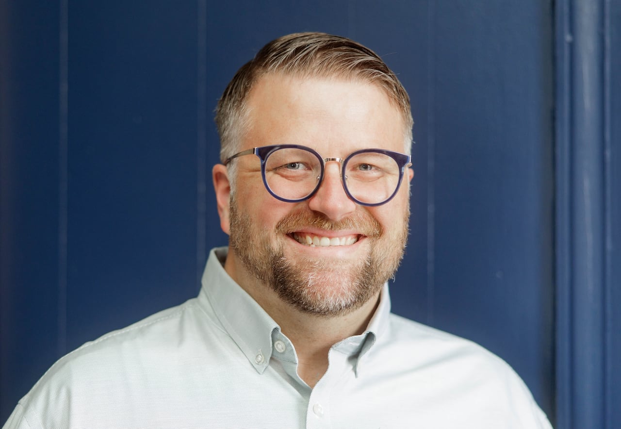 A white man with round glasses, light light blue eyes and light brown and grey hair and beard smiles in a headshot.
