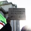 A protestor holds a cardboard sign in front of two Iranian flags. The sign reads "break communication blackout #freeIran".