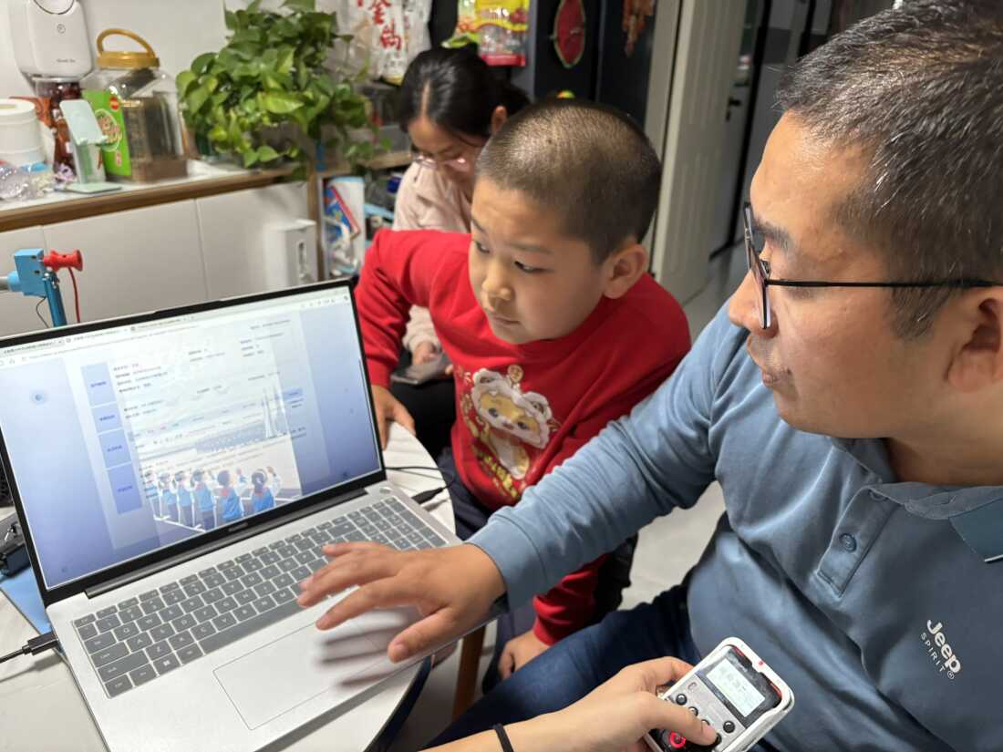 Li Zichen looks on as his father uses a computer in their home, with his mother in the background.
