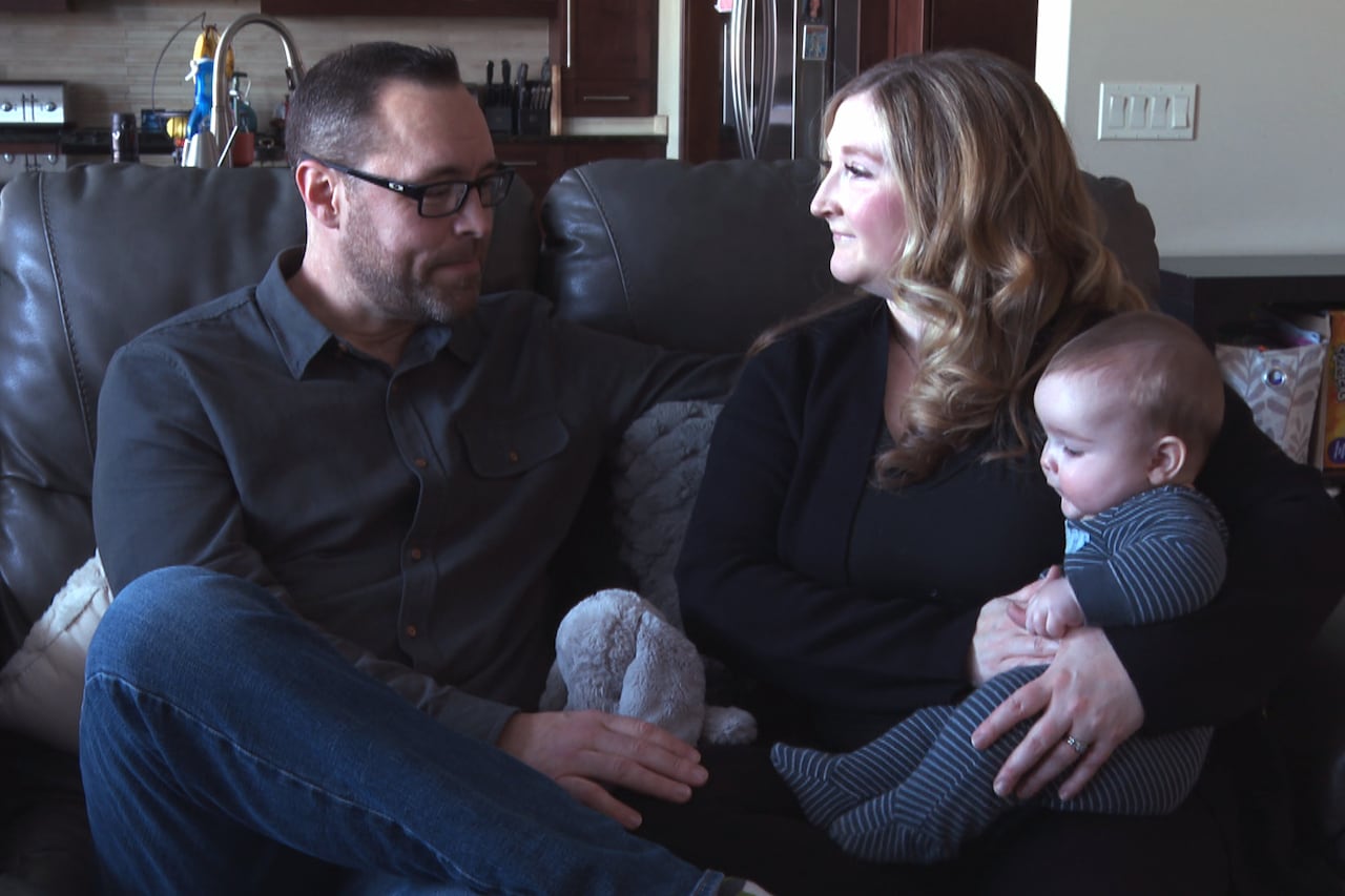 couple, male on left with baby on lap, women beside him on couch holding stuffed animal, kitchen behind them
