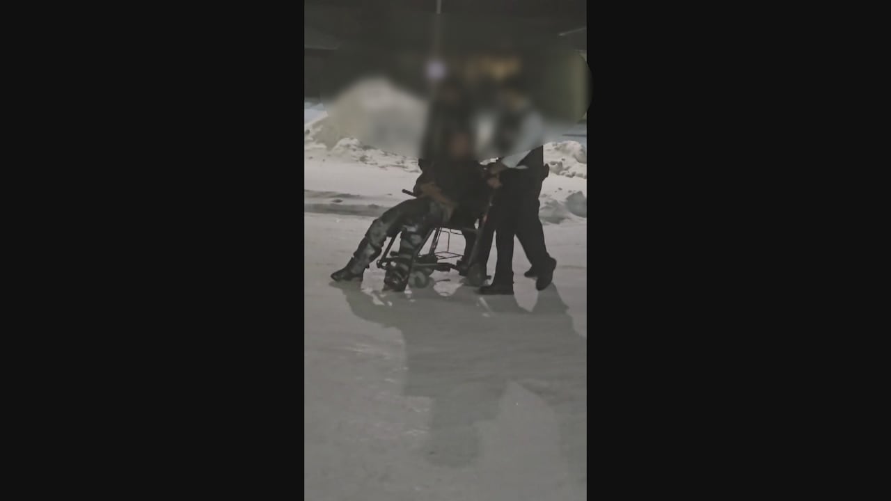 A blurry image of three men in dark clothing pushing a man in a wheelchair in a snowy parking lot.