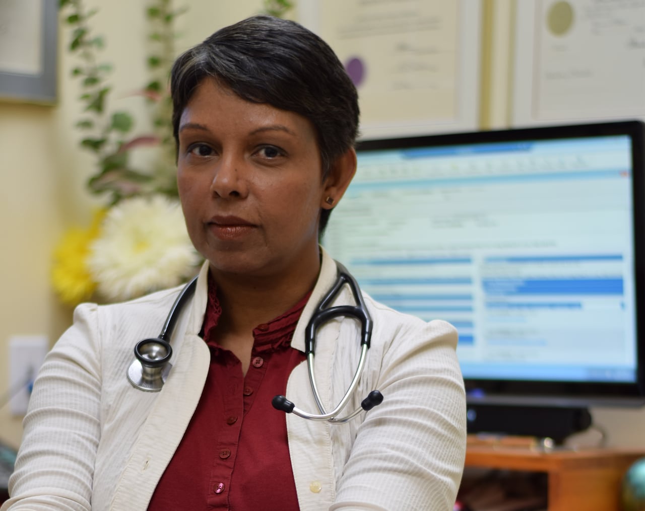 A short-haired woman in a lab coat with a stehoscope around her neck sits facing away from a computer