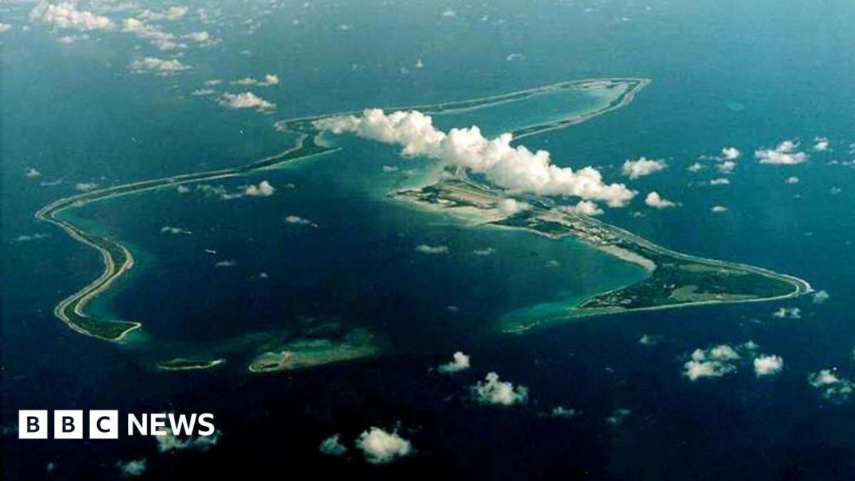 An aerial view of Diego Garcia, the largest island in the Chagos archipelago. Fluffy white clouds are above the island, which is surrounded by deep blue water.