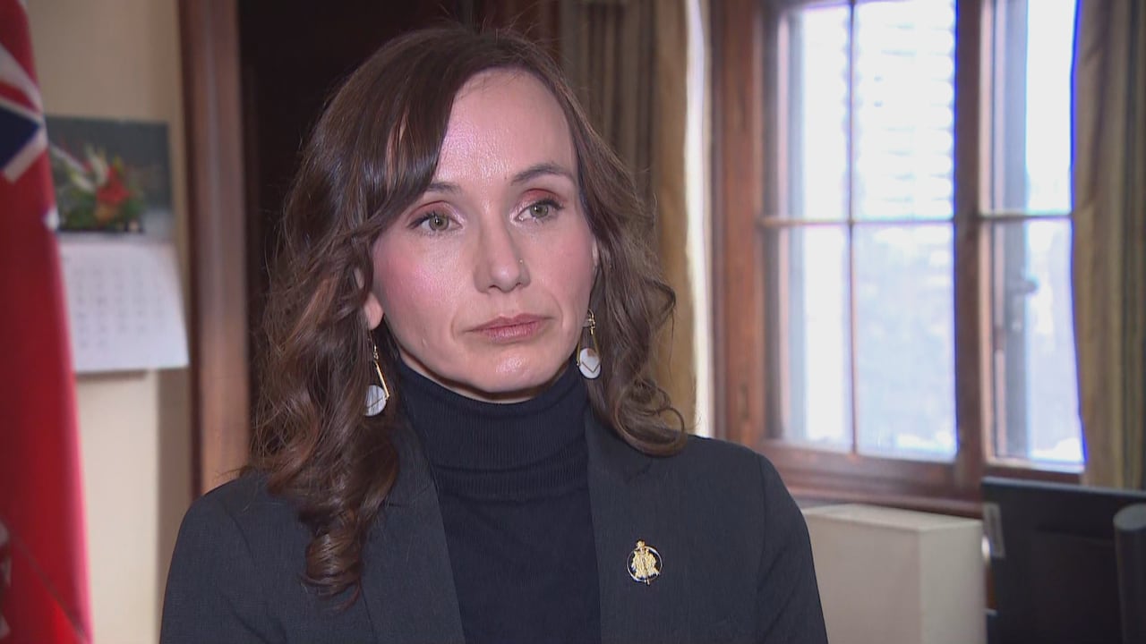 Woman with long  brown hair wears a dark blazer with a turtleneck shirt underneath and a gold pin on the lapel