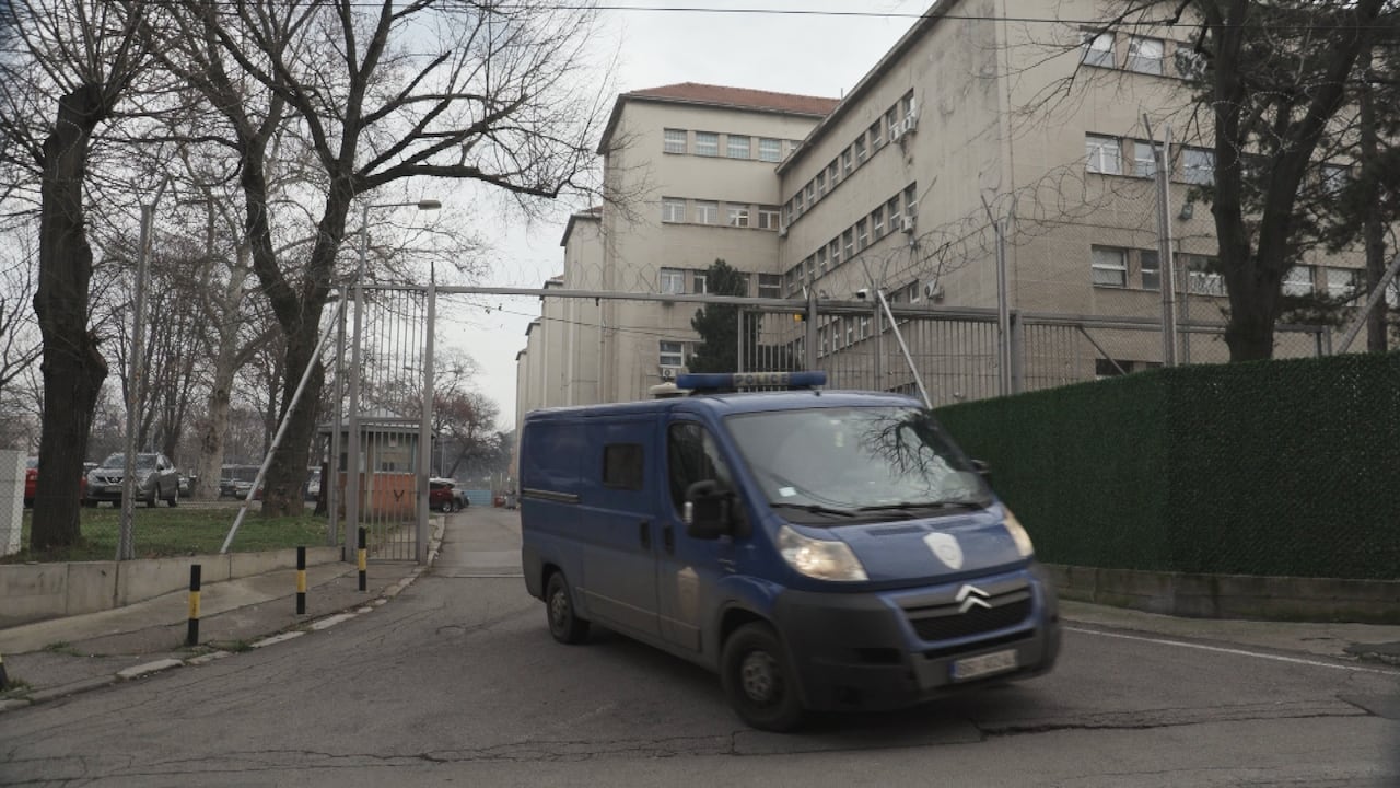 A blue van drives out of the high gate and fenced area surrounding the prison. 